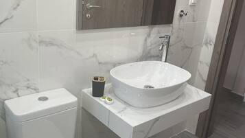 Un baño moderno con un inodoro blanco, un lavamanos grande y redondo, y un espejo sobre el lavamanos.