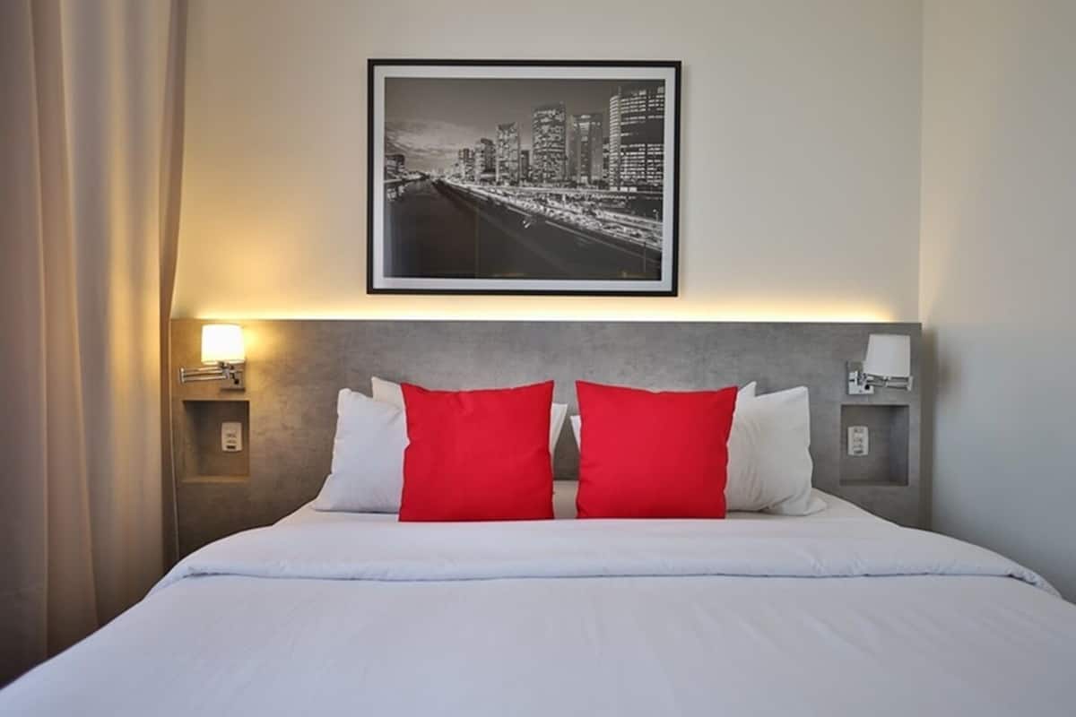 Une chambre d’hôtel avec un lit, deux oreillers rouges, un tableau représentant un paysage urbain encadré et des tables de chevet avec des lampes.