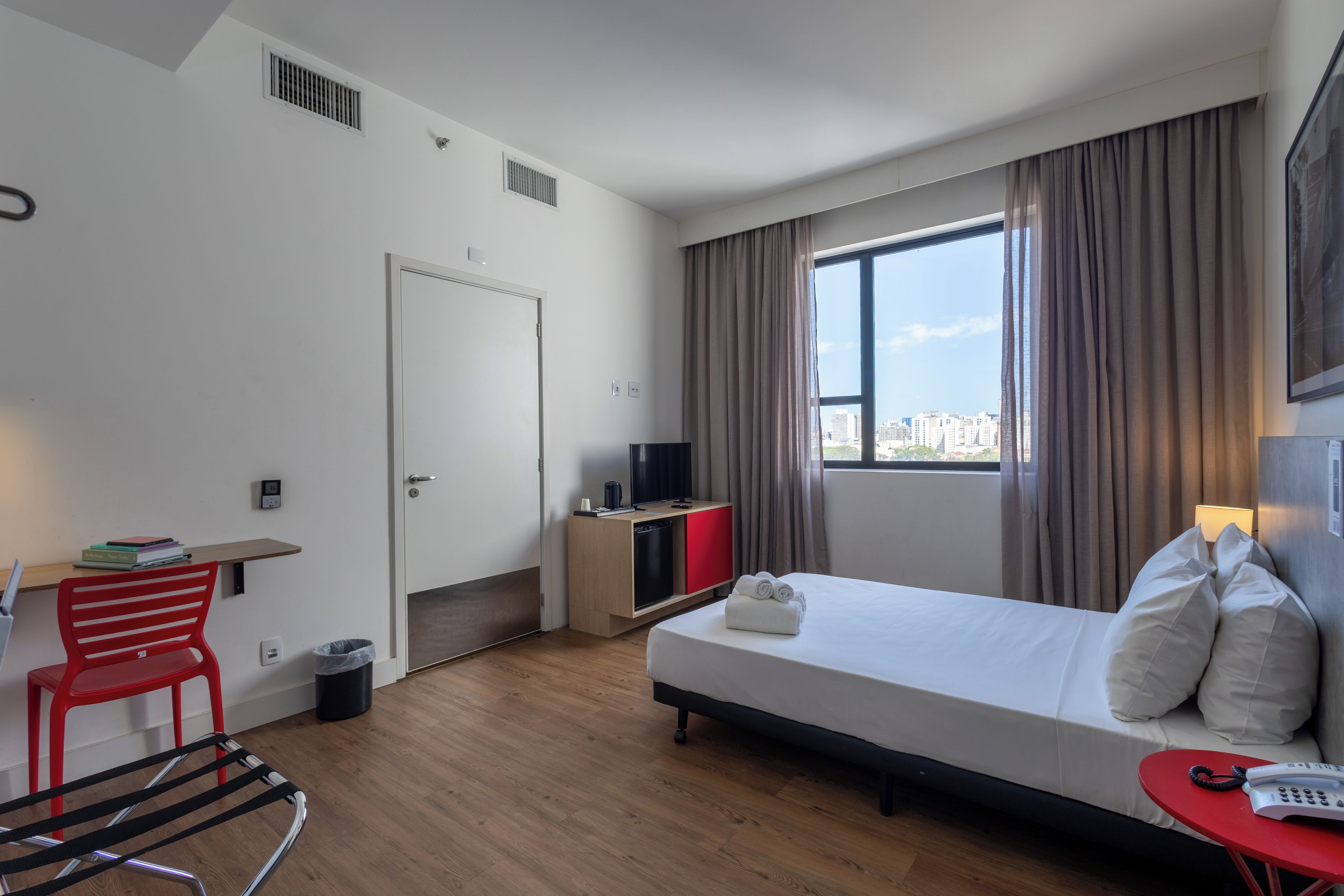Una habitación de hotel con cama, escritorio, silla, televisor y ventana con cortinas.