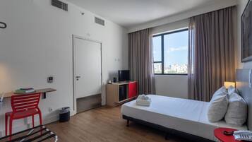 Una habitación de hotel con cama, escritorio, silla, televisor y ventana con cortinas.