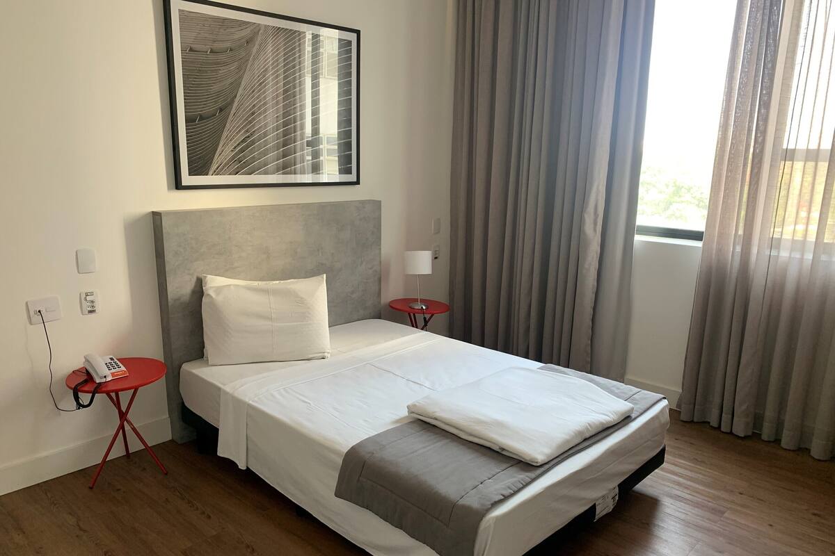 Un lit bien fait, recouvert d’une couverture grise, une table de chevet rouge avec un téléphone, et un tableau encadré accroché au mur.