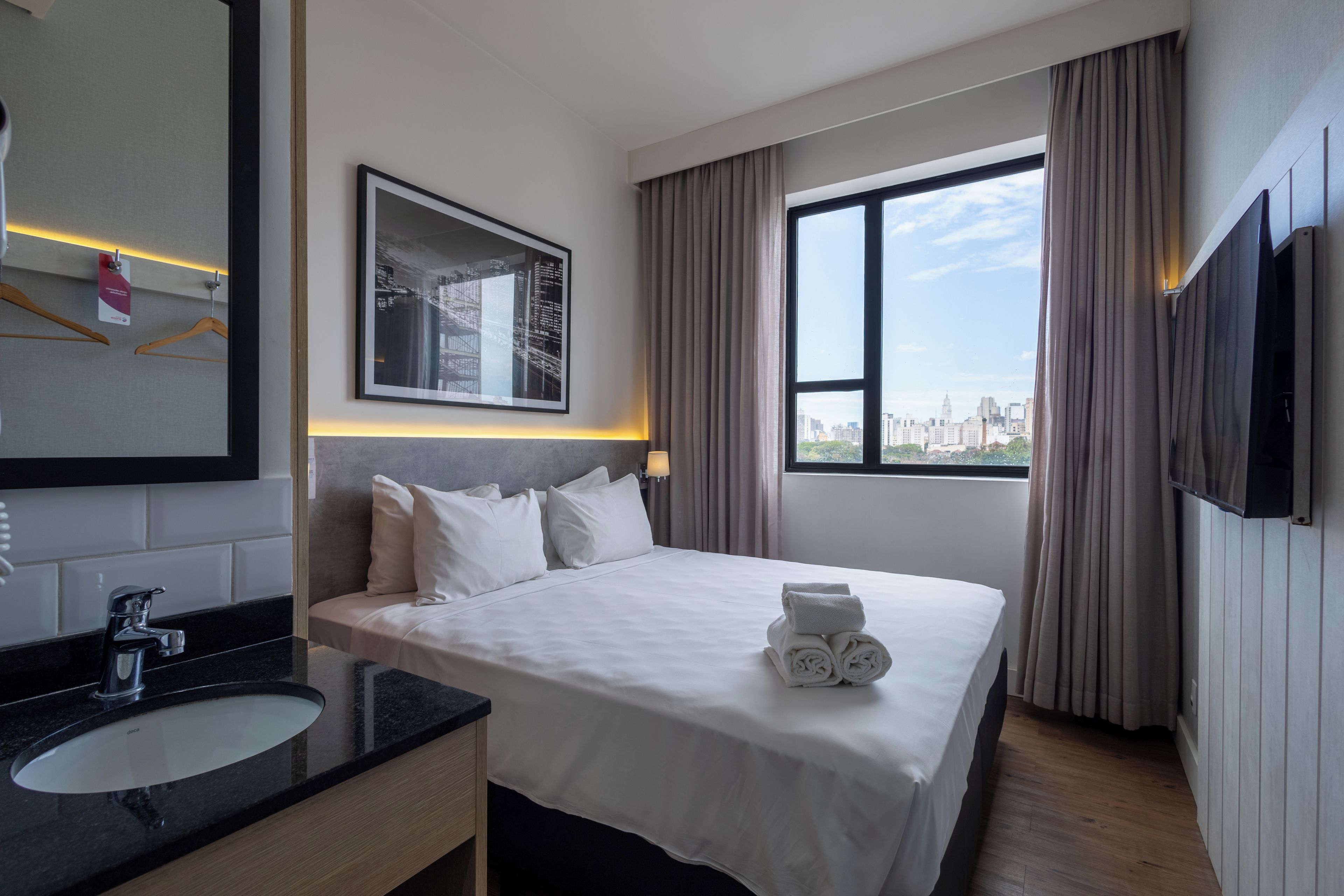 Une chambre d’hôtel avec un lit, une salle de bain comprenant un lavabo, un miroir et une fenêtre avec des rideaux.