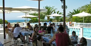 A poolside dining area with tables and umbrellas, overlooking a pool and the ocean. - Katerina Hotel Apartments (Preveza)