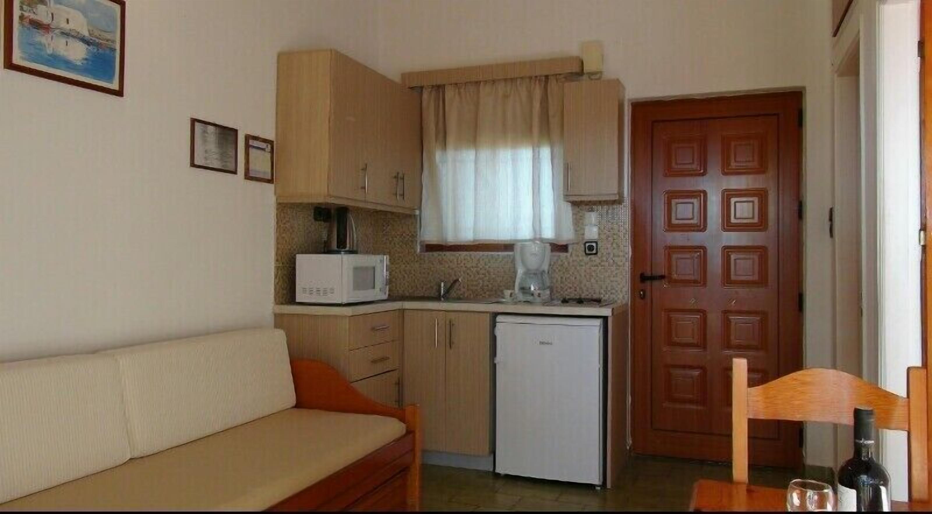A compact kitchenette with a microwave, coffee maker, and a small refrigerator. There is a wooden door with a square pattern, a beige sofa, and a small table with a bottle and glass.