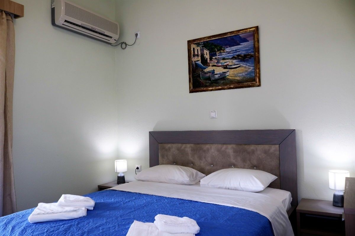 A hotel room with a bed, a painting, an air conditioner, and a nightstand with a lamp.