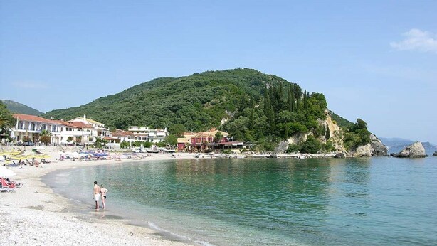 Una playa costera con edificios, agua cristalina y una colina al fondo.