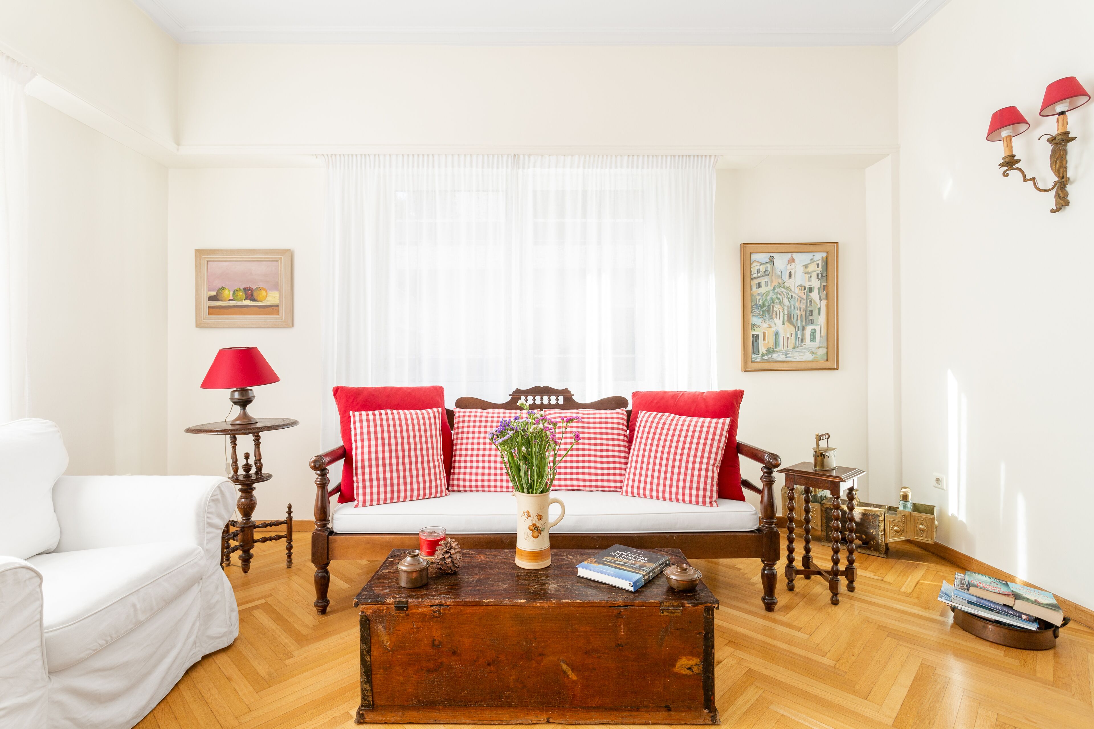 A living room with a white sofa, red and white checkered cushions, a wooden coffee table with a vase and books, and a red lamp.