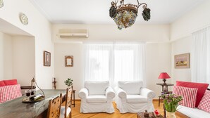 A living room with a wooden herringbone floor, white armchairs, a red and white checkered sofa, a wooden coffee table, and a chandelier. - Athens Squared Lofts (Athens)