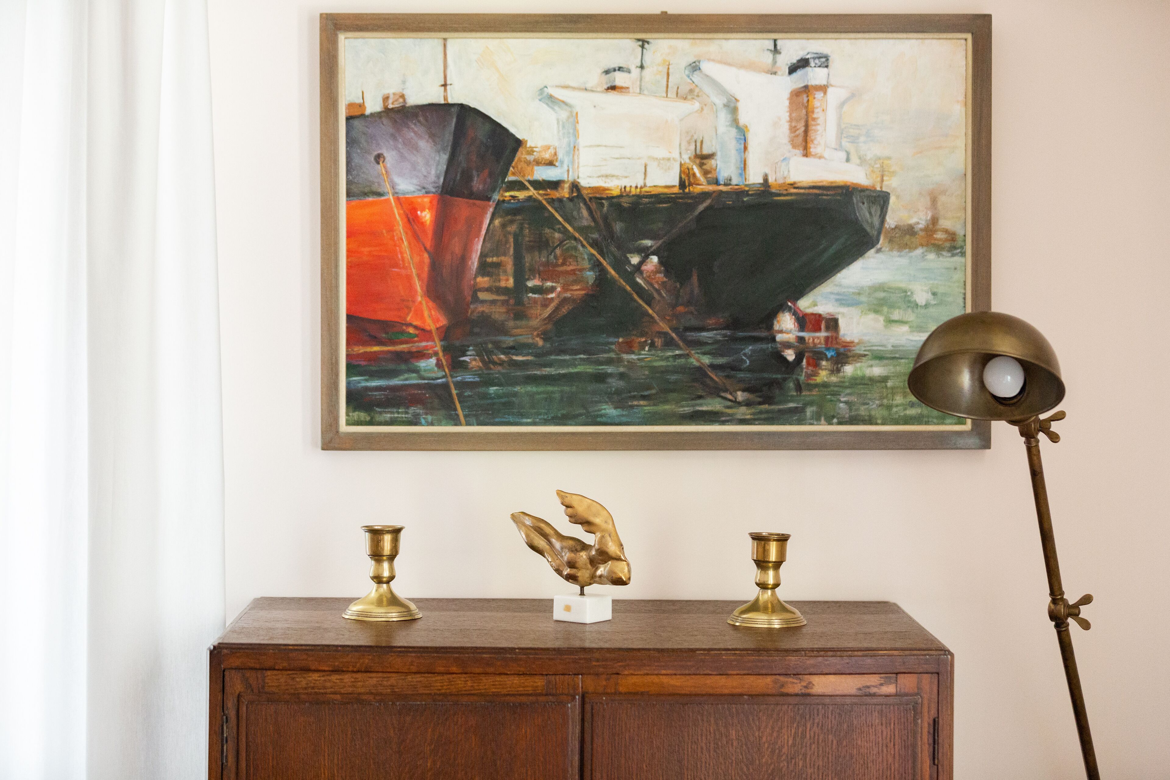 Un dipinto con navi su una parete, una scultura in ottone su un mobile in legno e una lampada da terra.