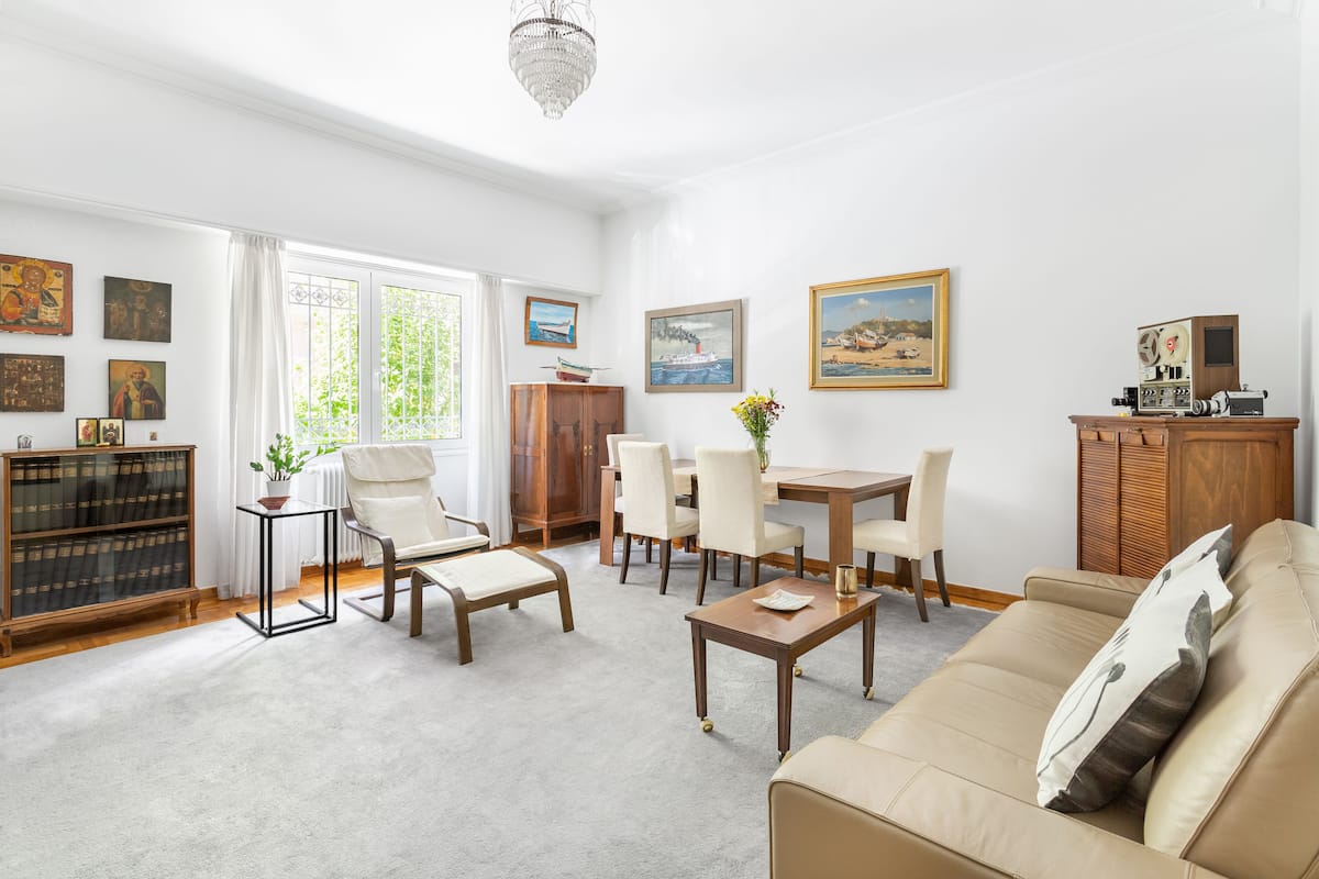 A spacious living room with a beige sofa, a dining table with chairs, a wooden cabinet, and various framed artworks on the walls.