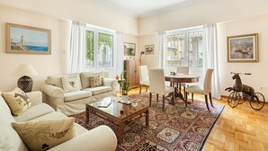 A spacious living room with a sofa set, a dining table, and chairs. There is a large window with curtains, a decorative rug, and a small table with a lamp. - Athens Squared Lofts (Athens)