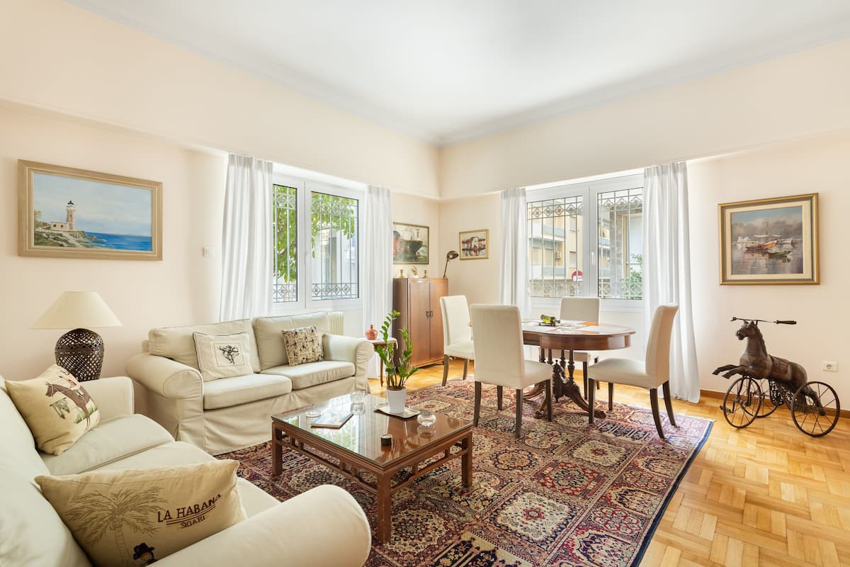 A spacious living room with a sofa set, a dining table, and chairs. There is a large window with curtains, a decorative rug, and a small table with a lamp.