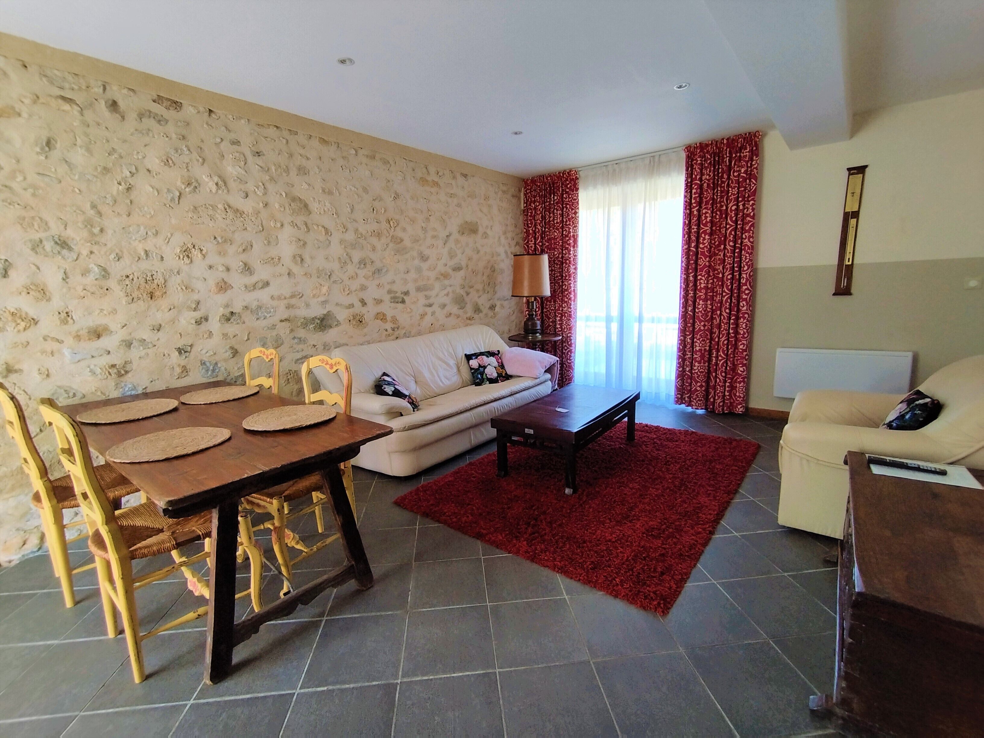 Een woonkamer met een stenen muur, een bank, een eettafel met stoelen, een rode vloerkleed en een lamp.
