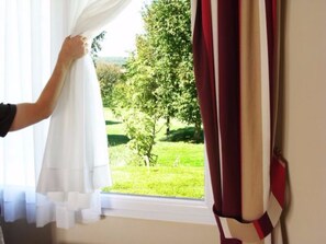 A window with white curtains partially drawn, revealing a view of a green landscape with trees and grass. - Le Haut-Val résidence (Belleme)