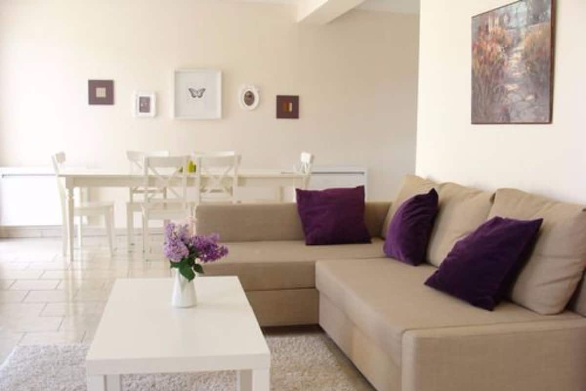 A modern living room with a beige sofa, white coffee table, and purple cushions. There is a dining area with a white table and chairs in the background.