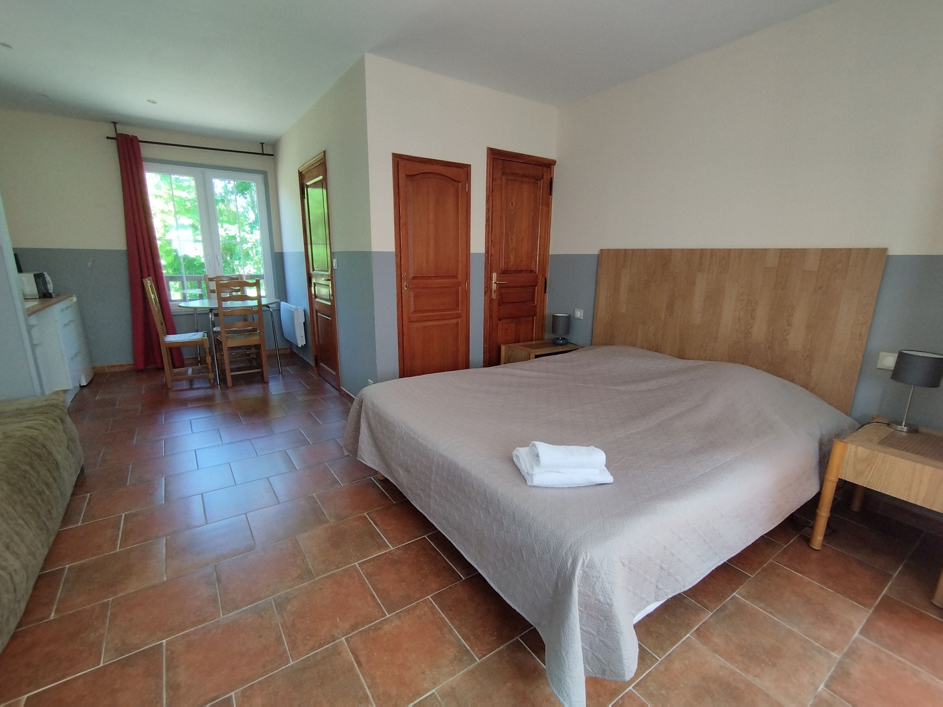 A bedroom with a large bed, a wooden headboard, a bedside table with a lamp, and a chair near a window.