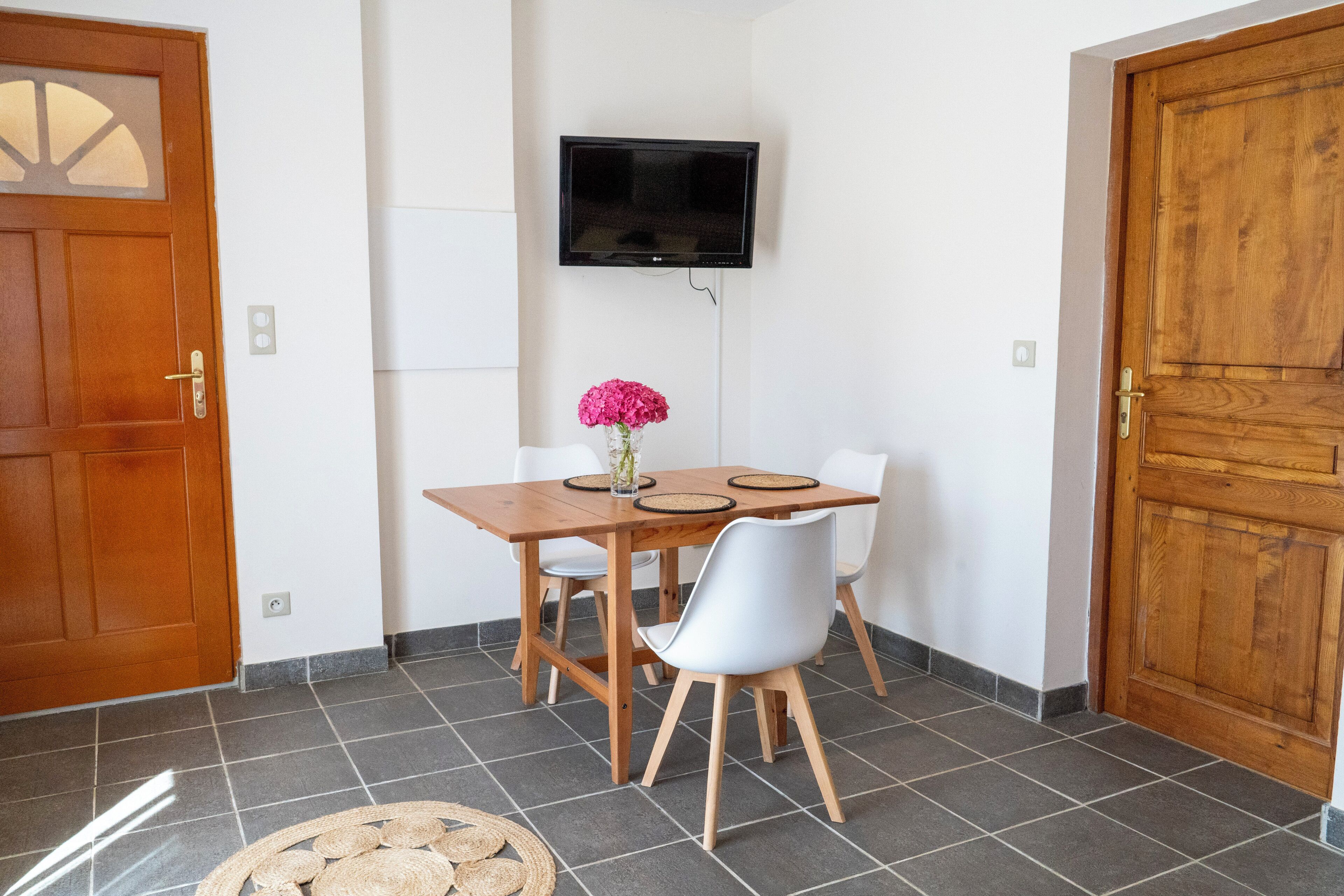 A dining area with a wooden table, white chairs, a TV mounted on the wall, and a wooden door.