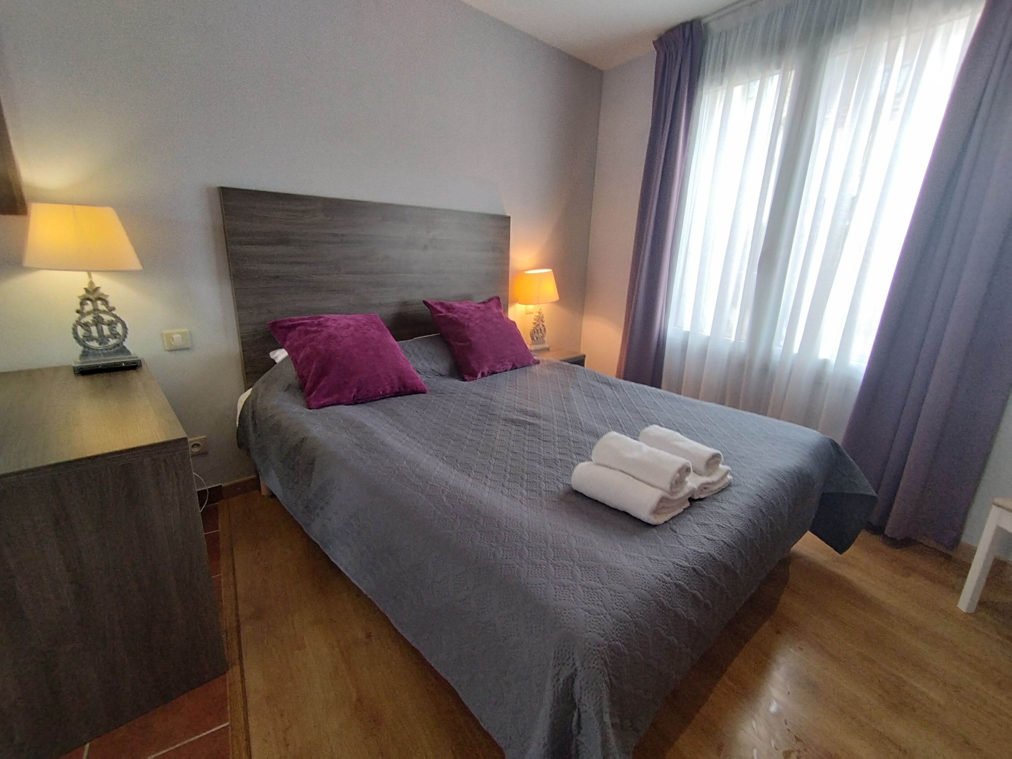 Un lit bien fait, recouvert d’une couverture grise et agrémenté de deux coussins violets, une table de chevet avec une lampe, et une fenêtre avec des rideaux.