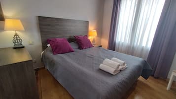 A neatly made bed with a grey bedspread and two purple pillows, a bedside table with a lamp, and a window with curtains.