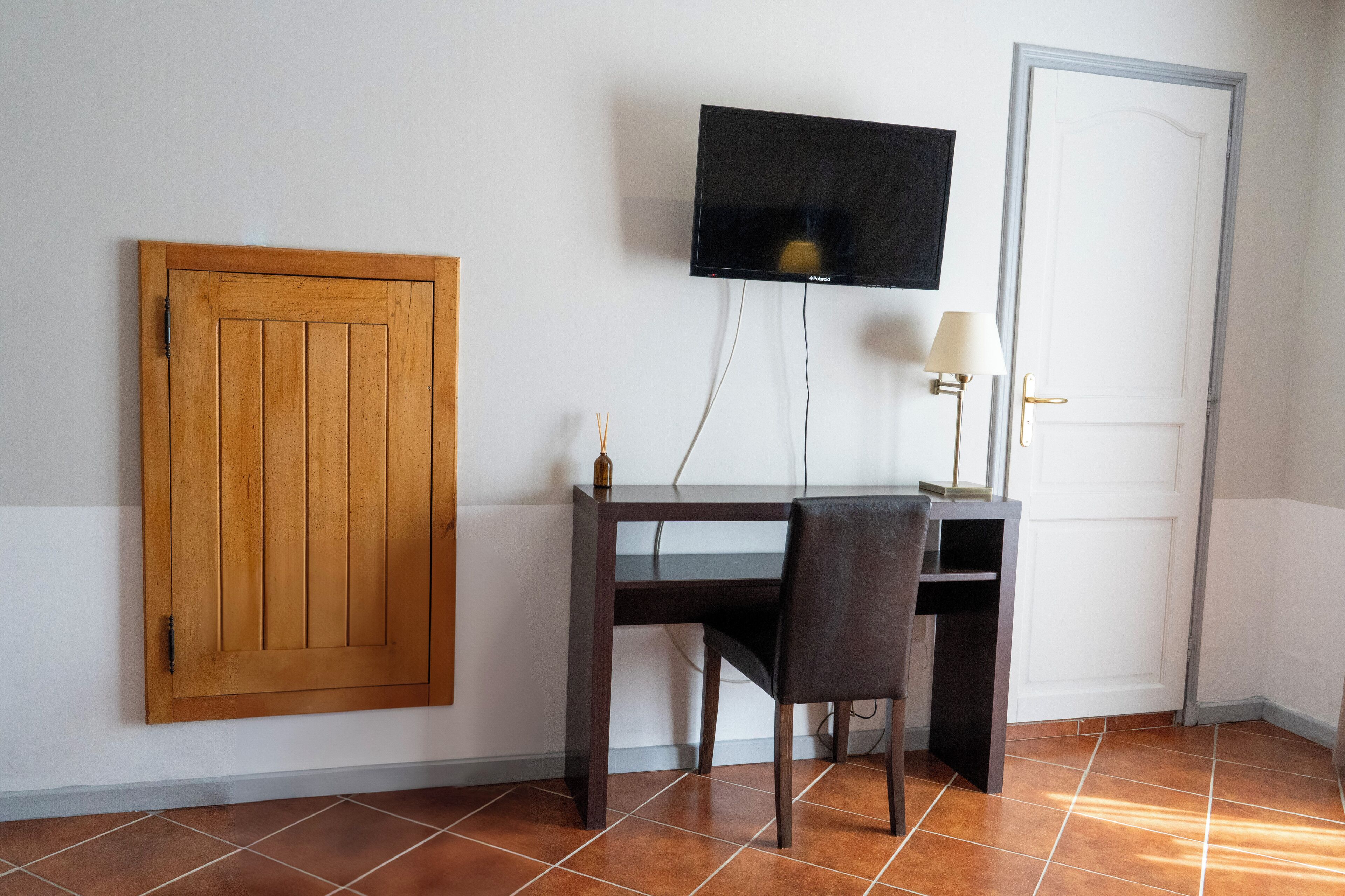 Ein Zimmer mit Holztür, Schreibtisch mit Stuhl, Wandfernseher und Lampe.