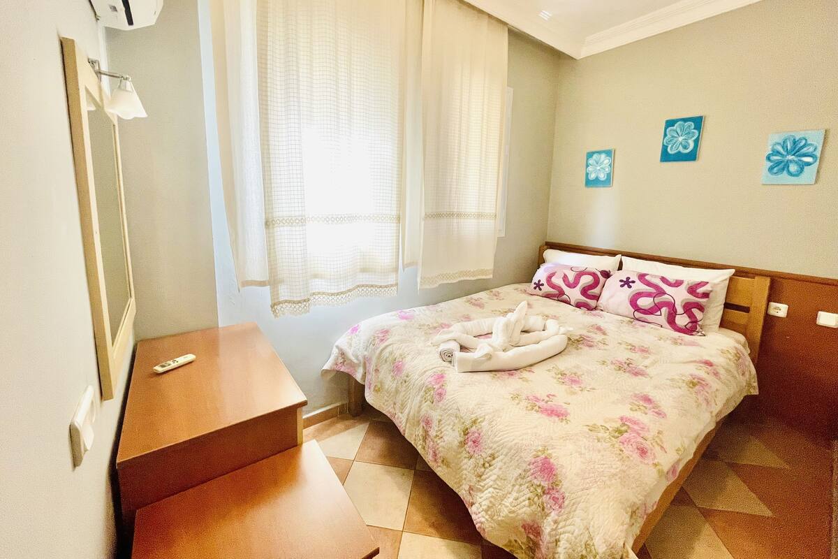A hotel room with a bed, a bedside table, a lamp, and a window with curtains.