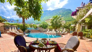 A poolside area with lounge chairs, a table with breakfast, and a view of mountains. - Mountain Valley Oludeniz (Fethiye)