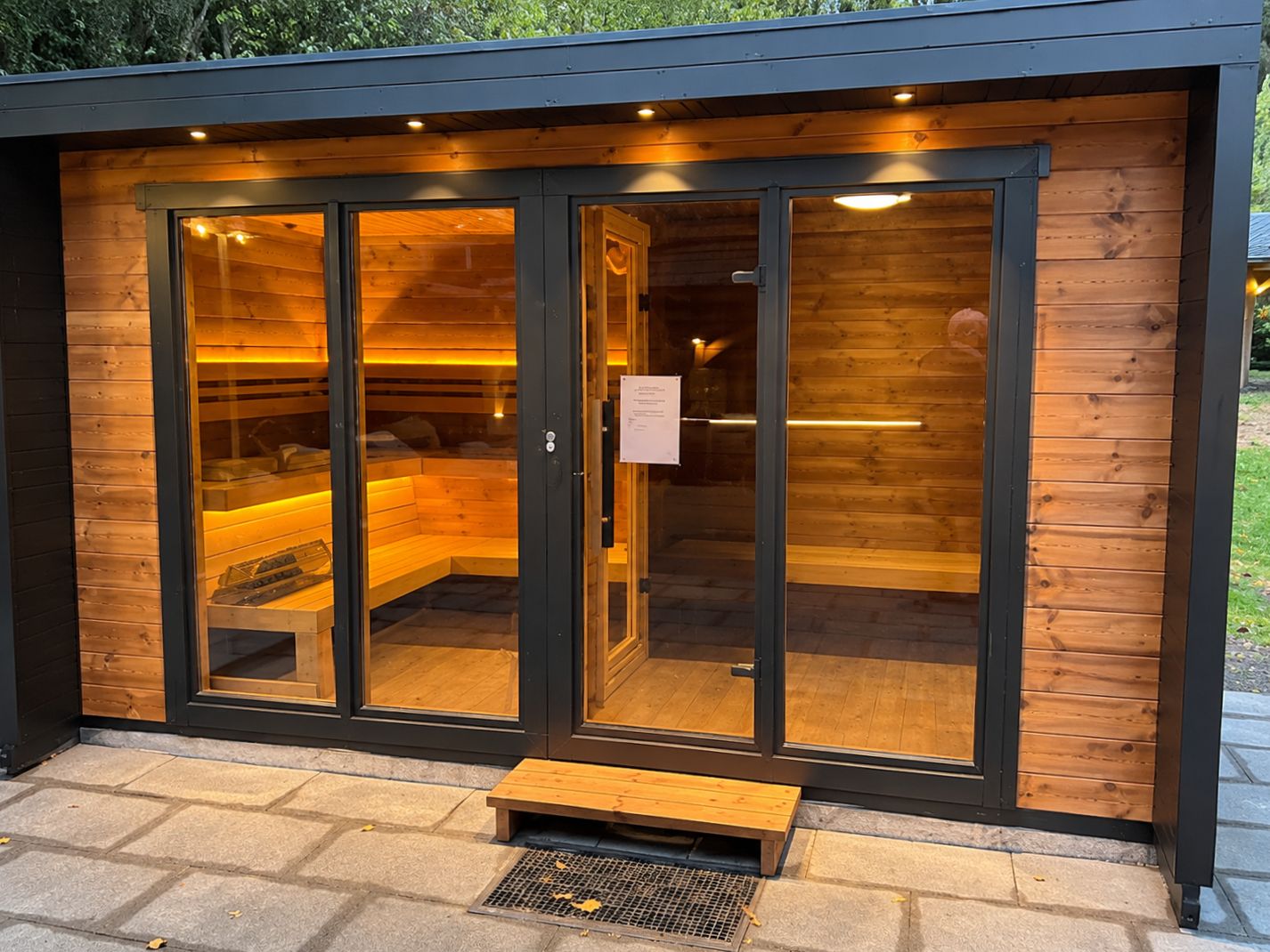 Eine Sauna mit Glastüren, einer Holzbank davor und einem Metallgitter.