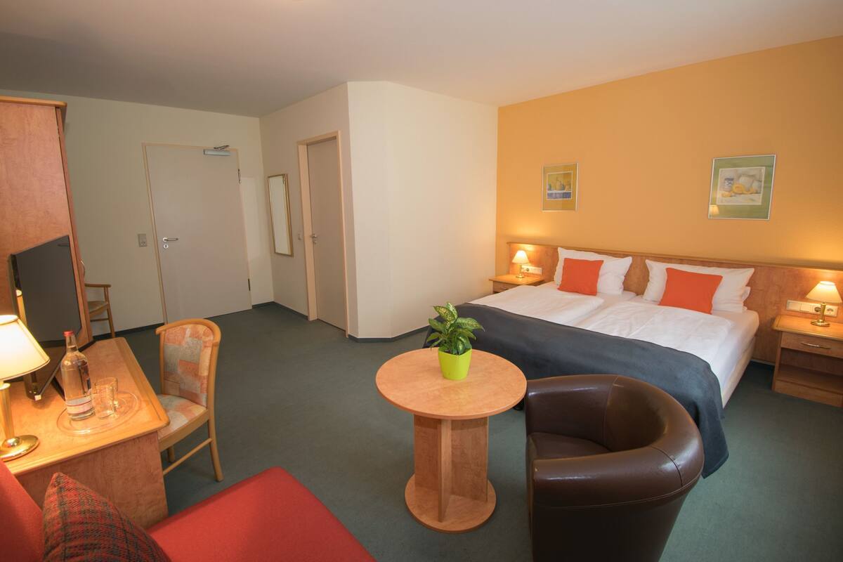 Een hotelkamer met een bed, een bureau, een stoel, een kleine tafel met een plant en een nachtkastje met een lamp.