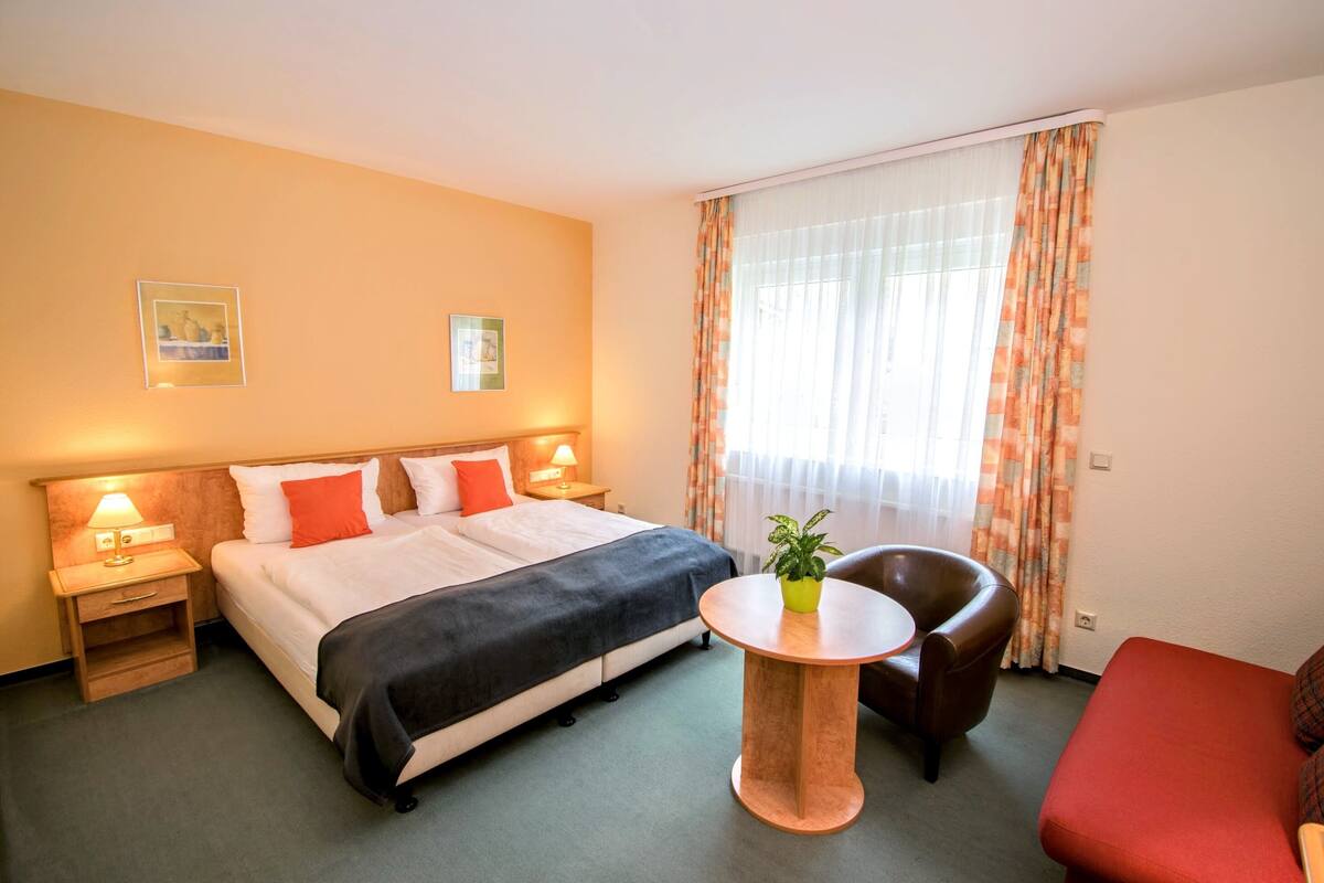 Een hotelkamer met een bed, nachtkastjes, een klein rond tafeltje met een plant, een leren fauteuil en een rode bank.