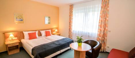 Habitación de hotel con cama, mesitas de noche, una pequeña mesa redonda con una planta, un sillón de cuero y un sofá rojo.