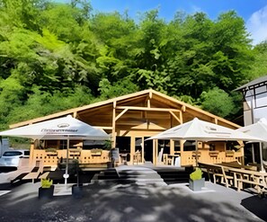 A wooden building with a pitched roof, surrounded by lush greenery. There are white umbrellas with logos on the ground in front. - Landhotel Ringelsteiner Mühle (Moselkern)