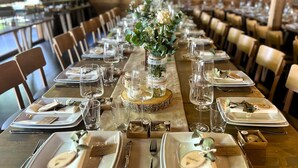 Een lange eettafel gedekt met borden, bestek en glazen, met een middelpunt van groen en bloemen.