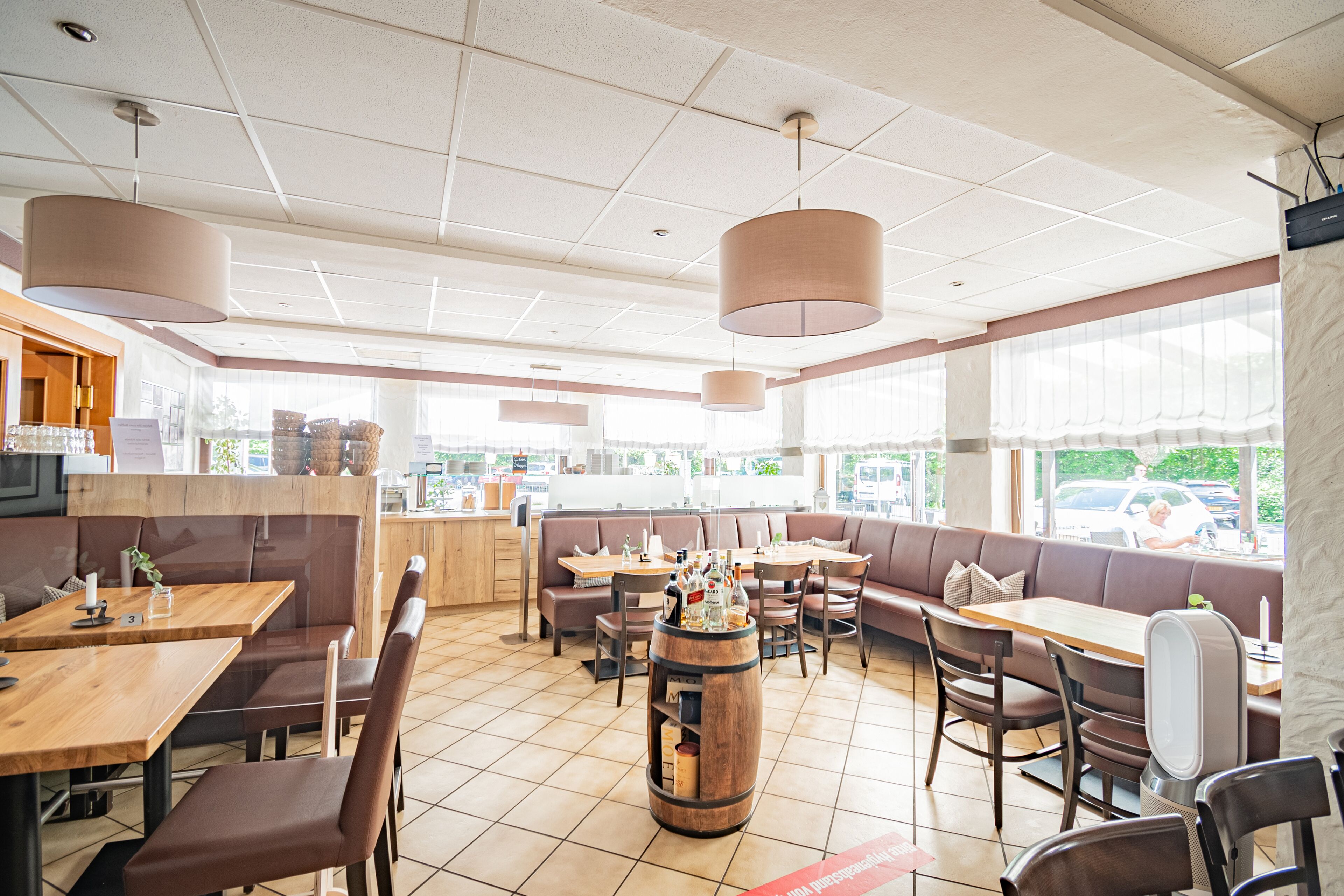 Un restaurant avec des tables et des banquettes en bois, un bar où sont exposées des bouteilles, et de grandes fenêtres qui laissent entrer la lumière naturelle.