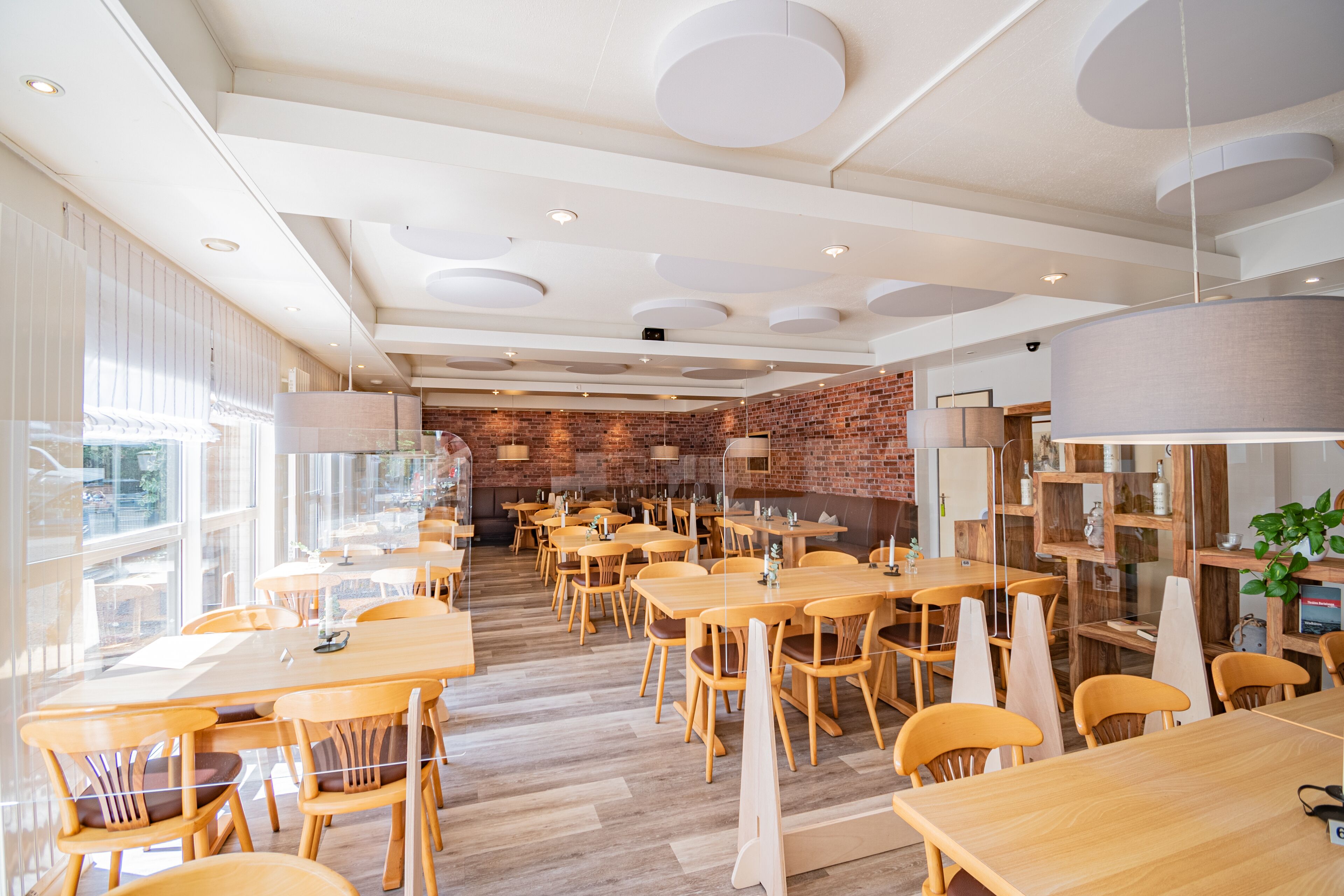 A spacious restaurant with wooden tables and chairs, a brick wall, and large windows.
