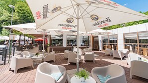 Een terras met witte stoelen en tafels, grote parasols met logo's, en een houten terrasvloer.