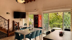 Un coin repas avec une table en verre, des chaises de couleur turquoise, un escalier en bois, un téléviseur et un réfrigérateur Coca-Cola.