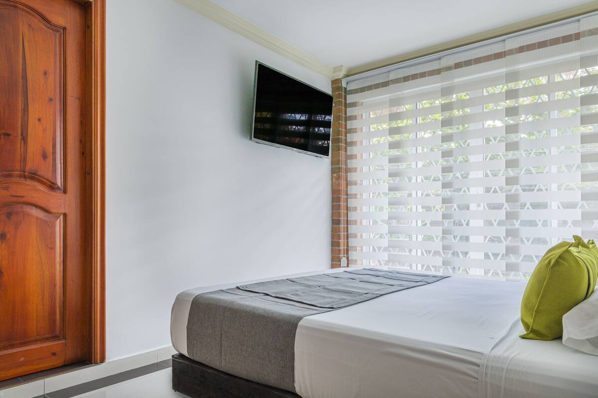 A bedroom with a bed, a television mounted on the wall, and a window with blinds.