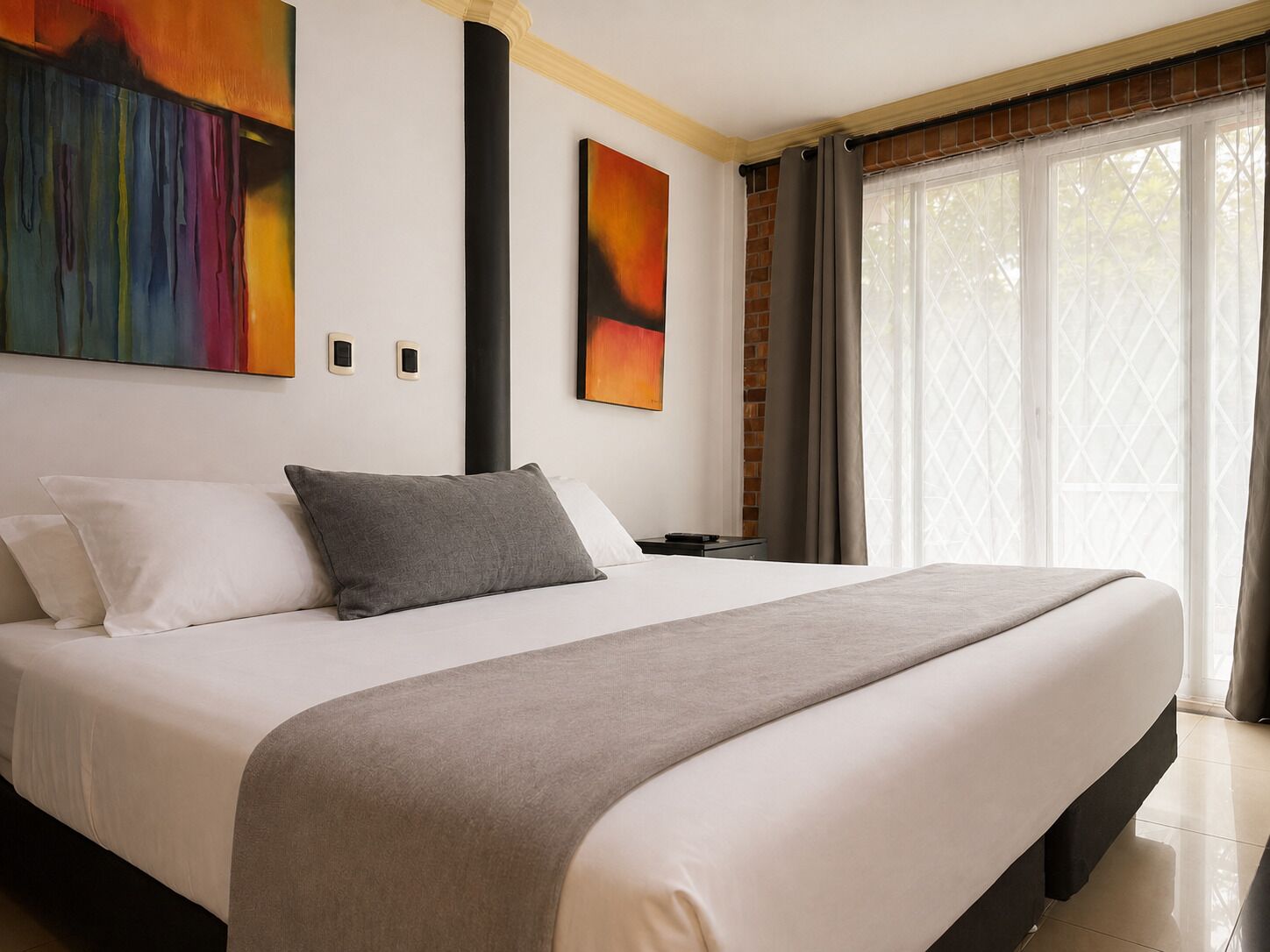 Quarto de hotel moderno com uma cama grande, uma mesa de cabeceira, uma luminária e uma janela com cortinas.