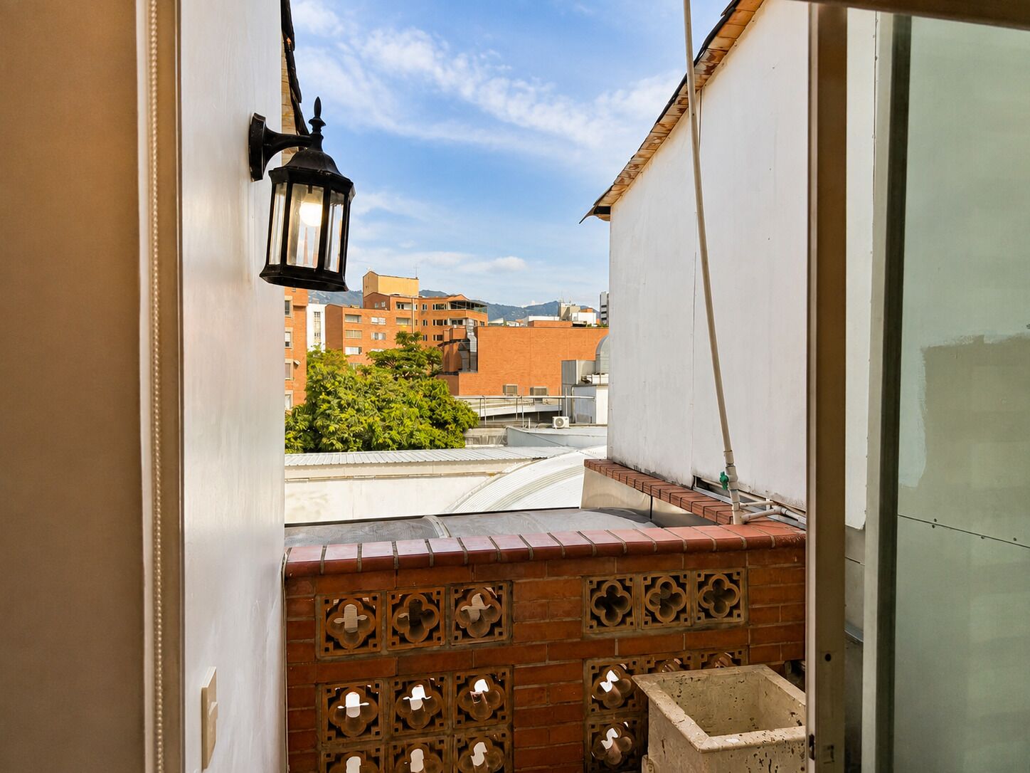Un balcón con un muro de ladrillo, un farol colgante y vistas a edificios y árboles.