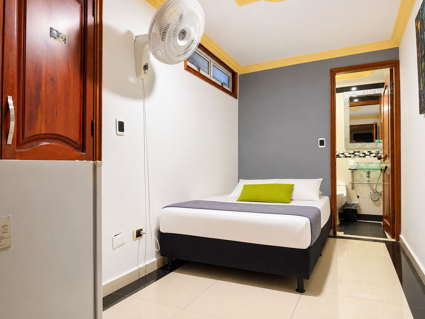 Un dormitorio con cama, ventilador de pared, armario de madera y baño visible a través de una puerta abierta.