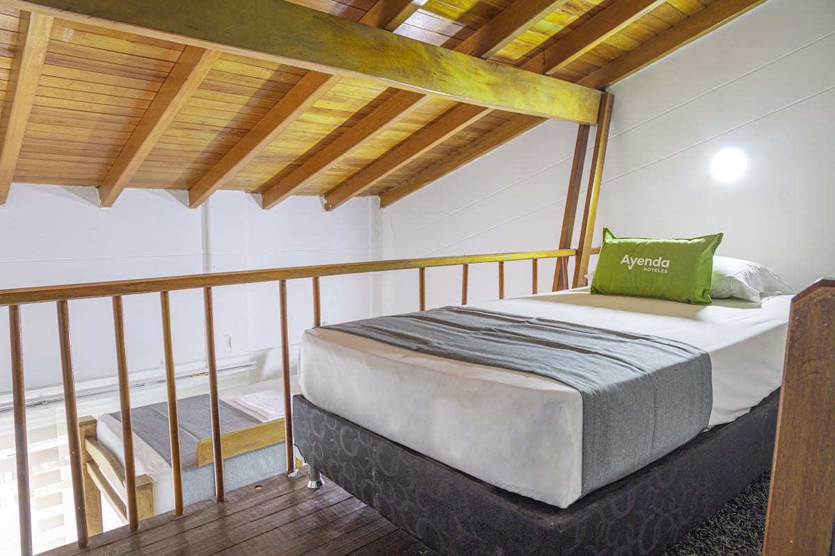 A bedroom with a wooden ceiling, a bed with a grey mattress and a green pillow, and a white wall.