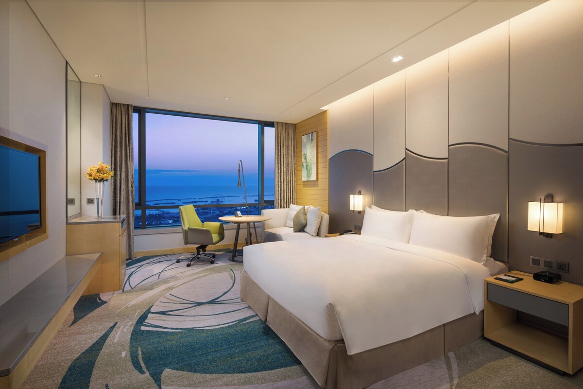 Habitación de hotel con una cama grande, un escritorio, una silla, una mesita y vistas al océano.