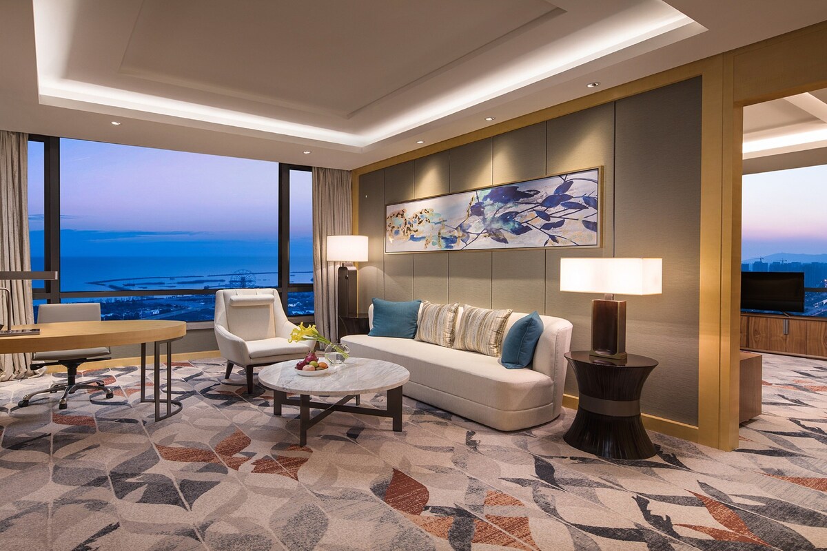 Habitación de hotel moderna con un amplio ventanal que ofrece vistas a la ciudad, un sofá, una mesita de centro y un televisor.