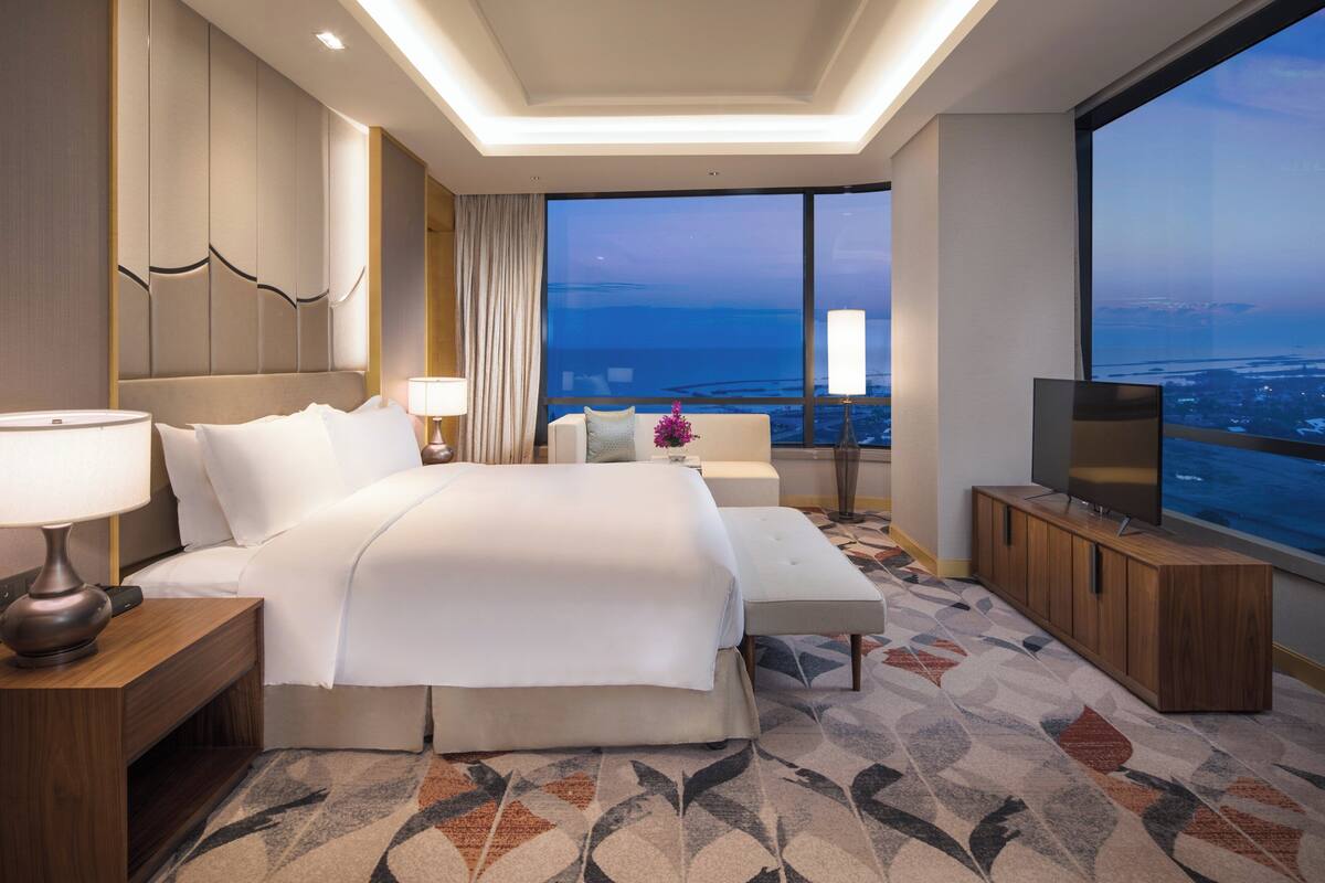 A modern hotel room with a large bed, a sofa, a TV, and a view of the cityscape.