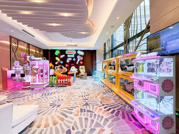 Zona luminosa de arcade con máquinas de garra y letreros de neón, con piso estampado y ventanas amplias.