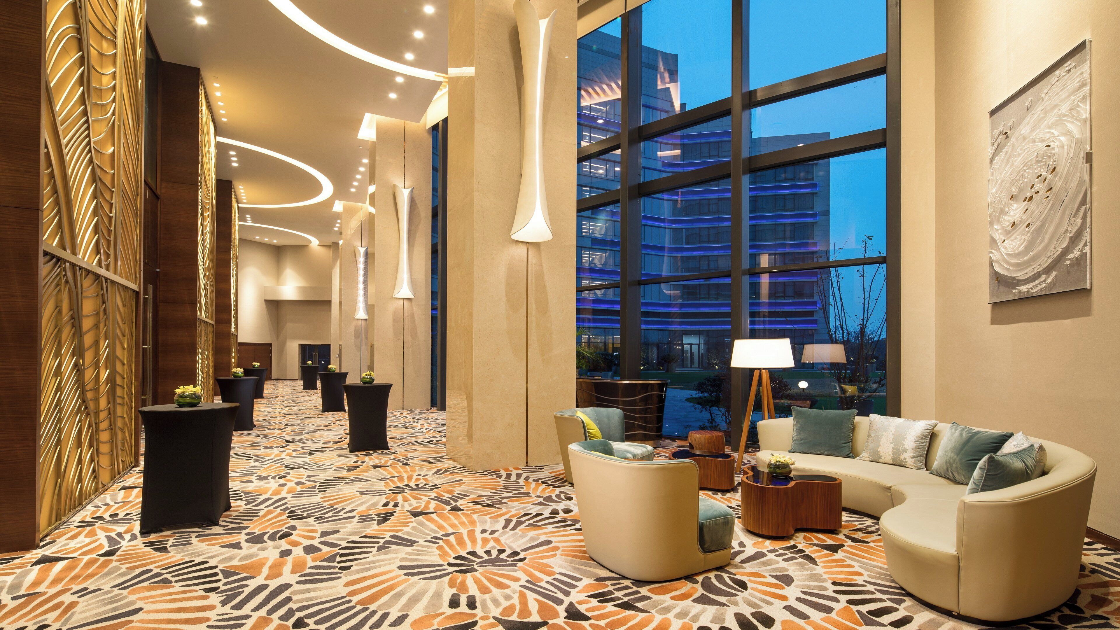 Un vestíbulo de hotel con alfombra estampada, muebles modernos y amplias ventanas que ofrecen vistas de un edificio.