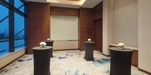 Un salón de conferencias con una pantalla grande, tres mesas negras con flores blancas y una alfombra estampada.