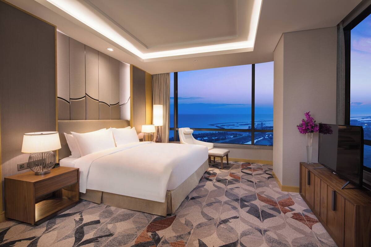 A modern hotel room with a large bed, a TV, a desk with a lamp, and a view of the ocean.