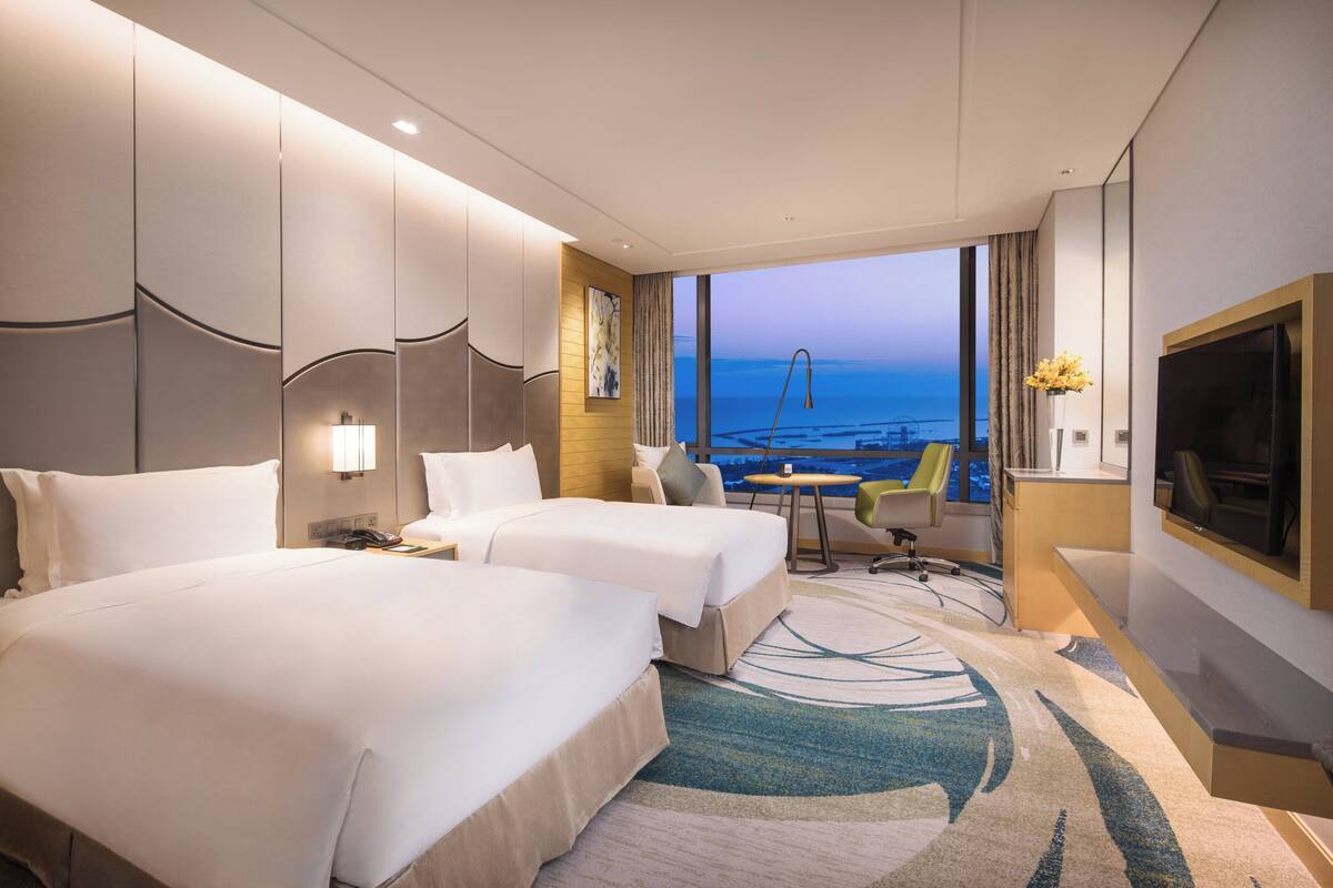 A hotel room with two beds, a TV, a desk, and a view of the ocean.