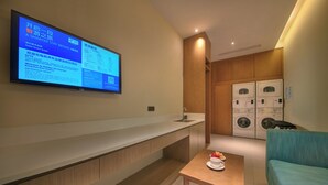 A modern hotel room with a flat-screen TV mounted on the wall displaying a hotel guide. There is a wooden bench along the wall, a small table with a plate of fruit, and a blue sofa. A washing machine is visible in the background. - Holiday Inn Express Hangzhou East Station by IHG (Hangzhou)